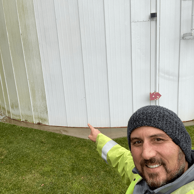 Man pointing at a building with green algae, wearing a beanie and safety vest.