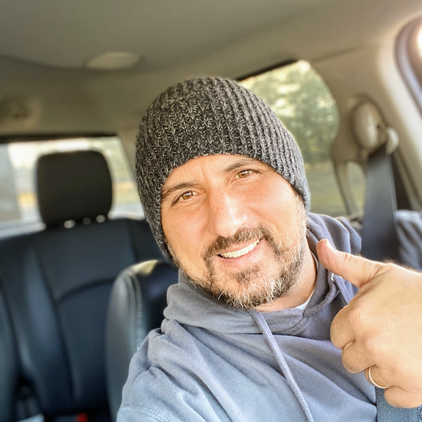 Man in gray beanie and hoodie gives a thumbs up from inside a car.