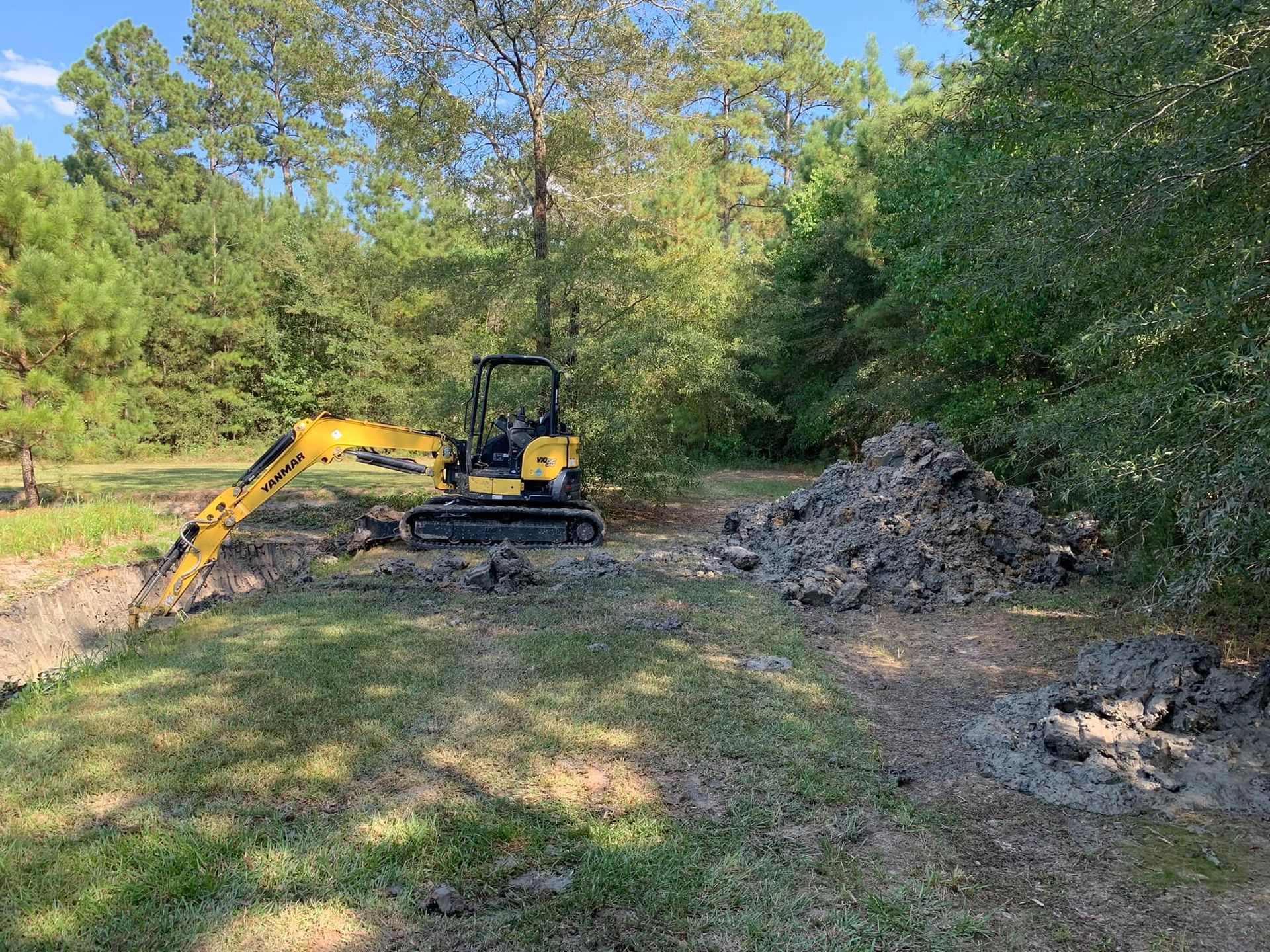 Yellow excavator digging in a grassy area, creating a trench. Dirt pile on the right, trees in the background.