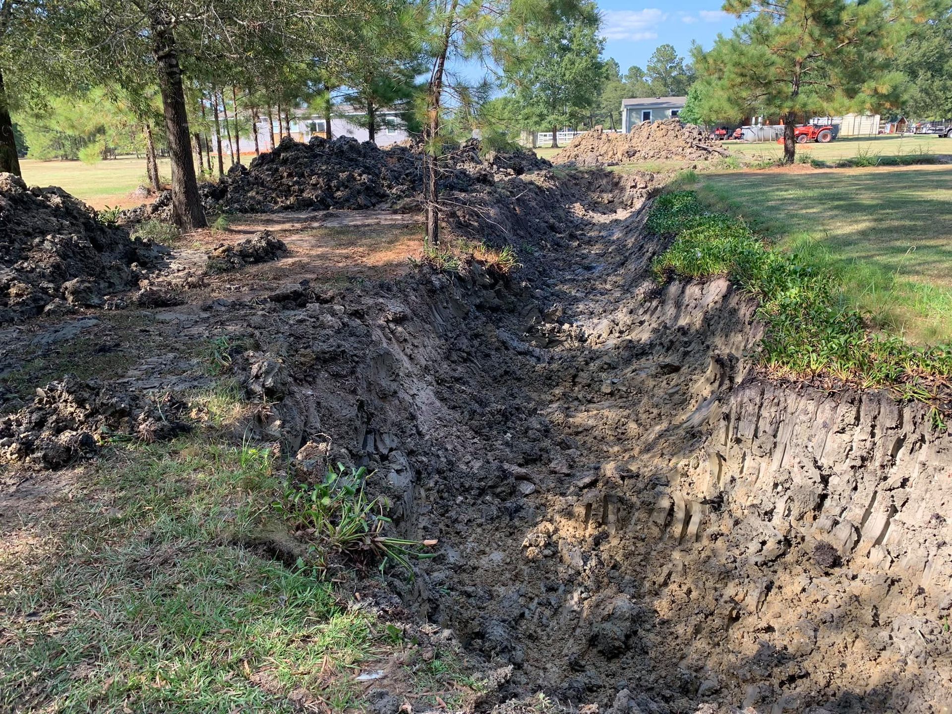 A muddy ditch dug in grassy land with piles of dirt nearby, trees in the background.