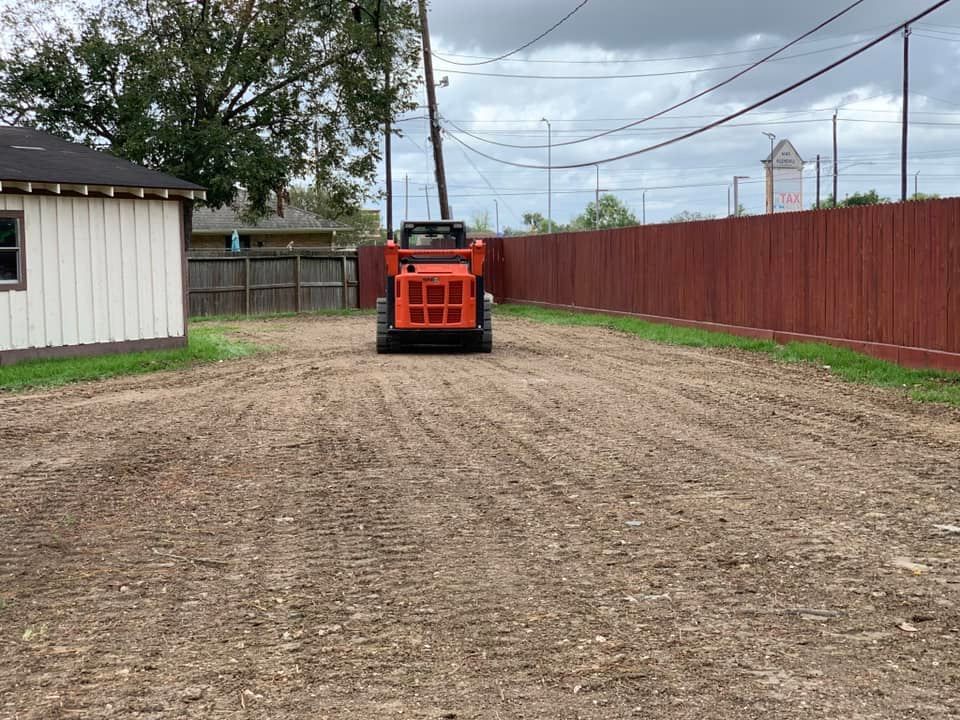An orange skid steer compacting dirt in a cleared lot, a red fence to the right and a small building to the left.