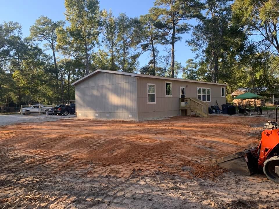 A single-story, beige mobile home on a leveled dirt lot with construction equipment nearby, trees in the background.