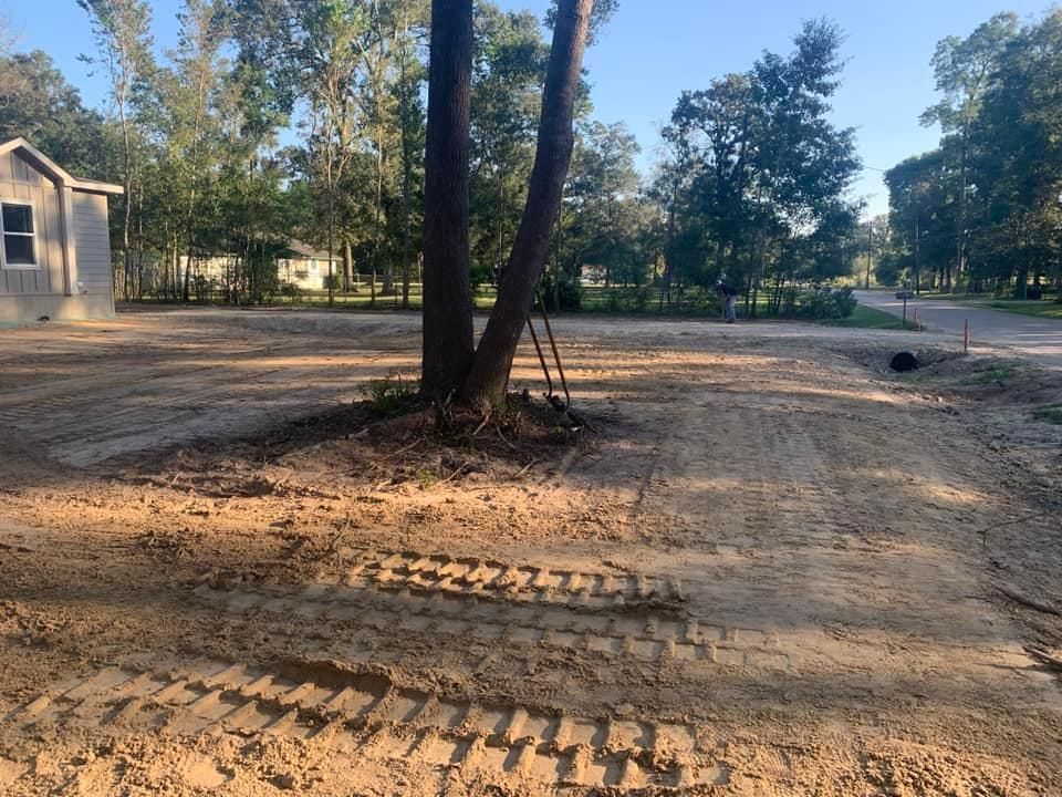 Dirt lot with tire tracks, trees in the center, and a house in the background.