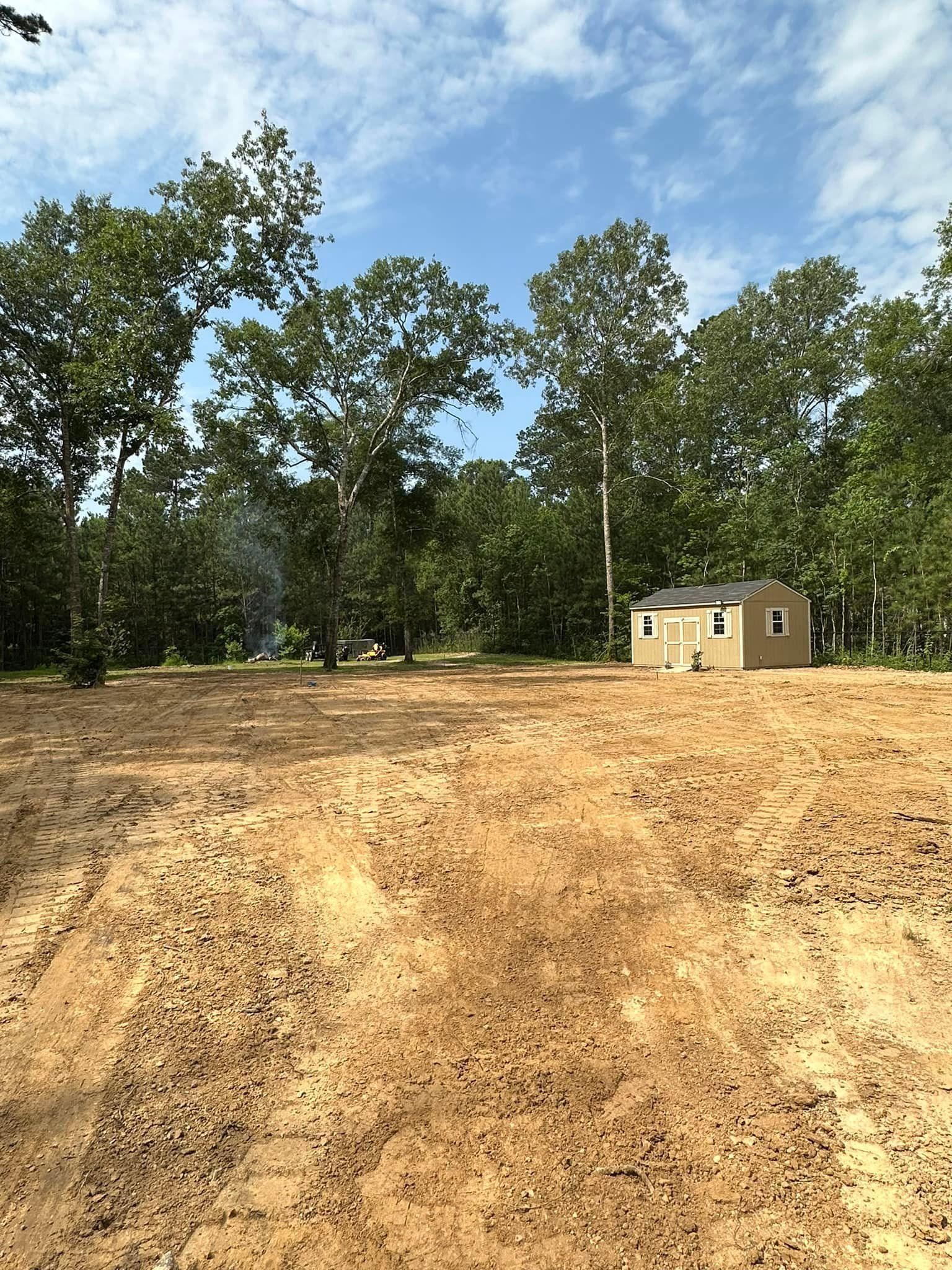 A cleared, dirt lot with a small shed, surrounded by trees under a blue sky.
