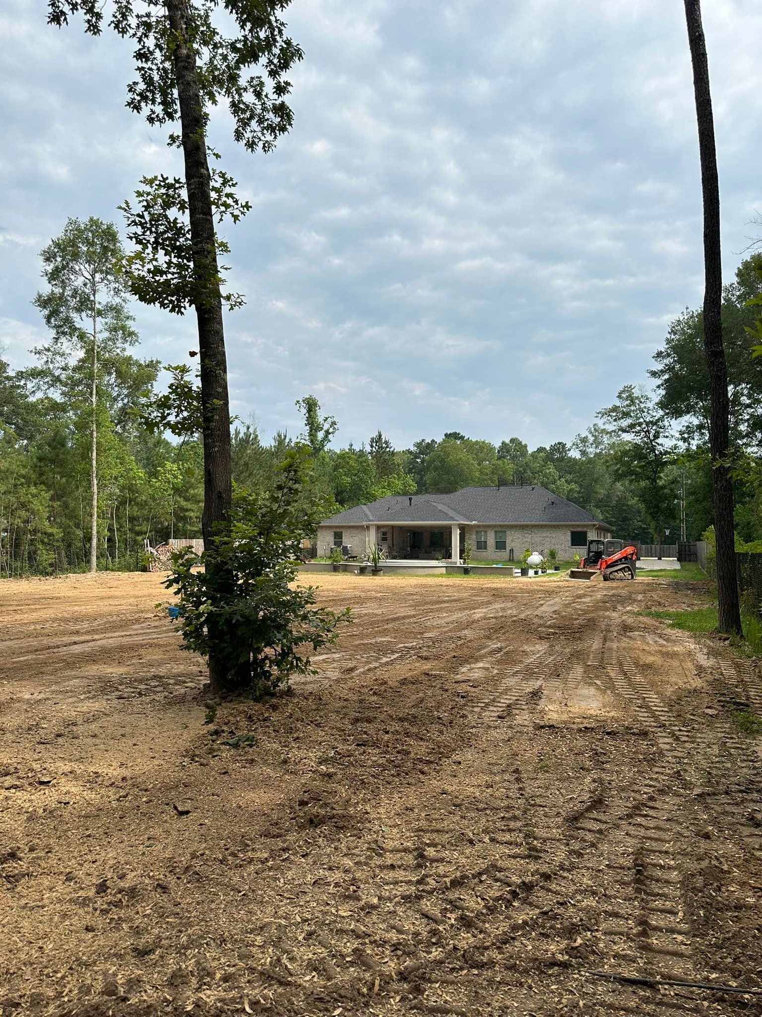 Dirt field with house in the background and trees on the left and right sides. Cloudy sky above.