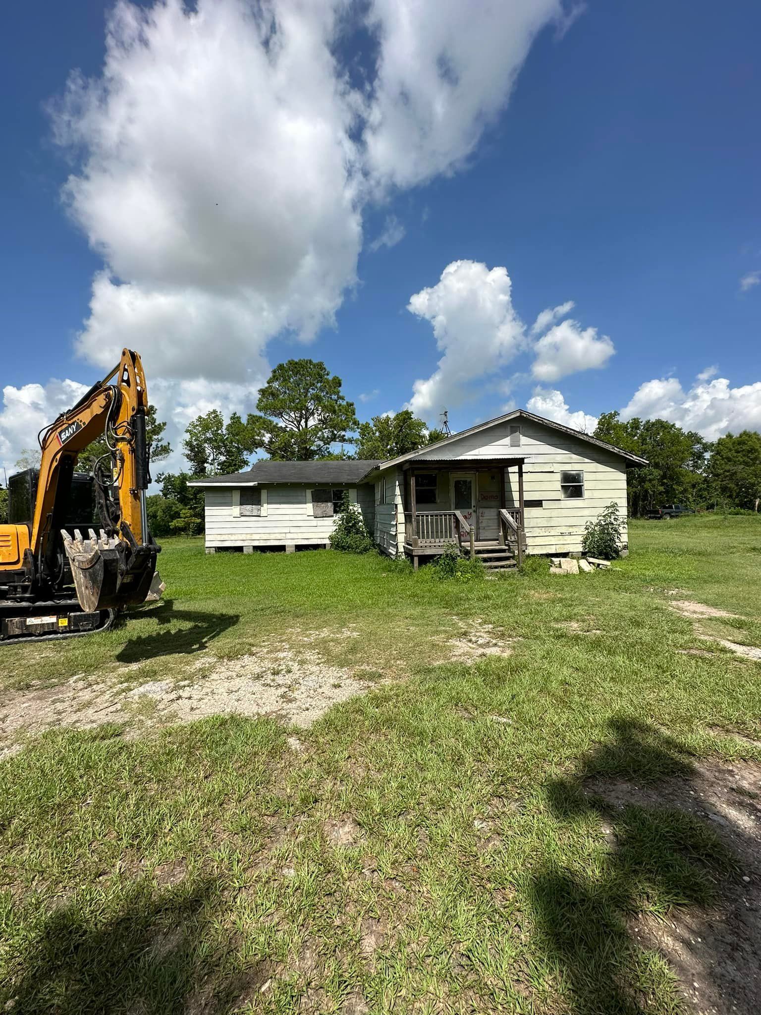 Old, weathered house with an excavator nearby on a sunny, grassy field.