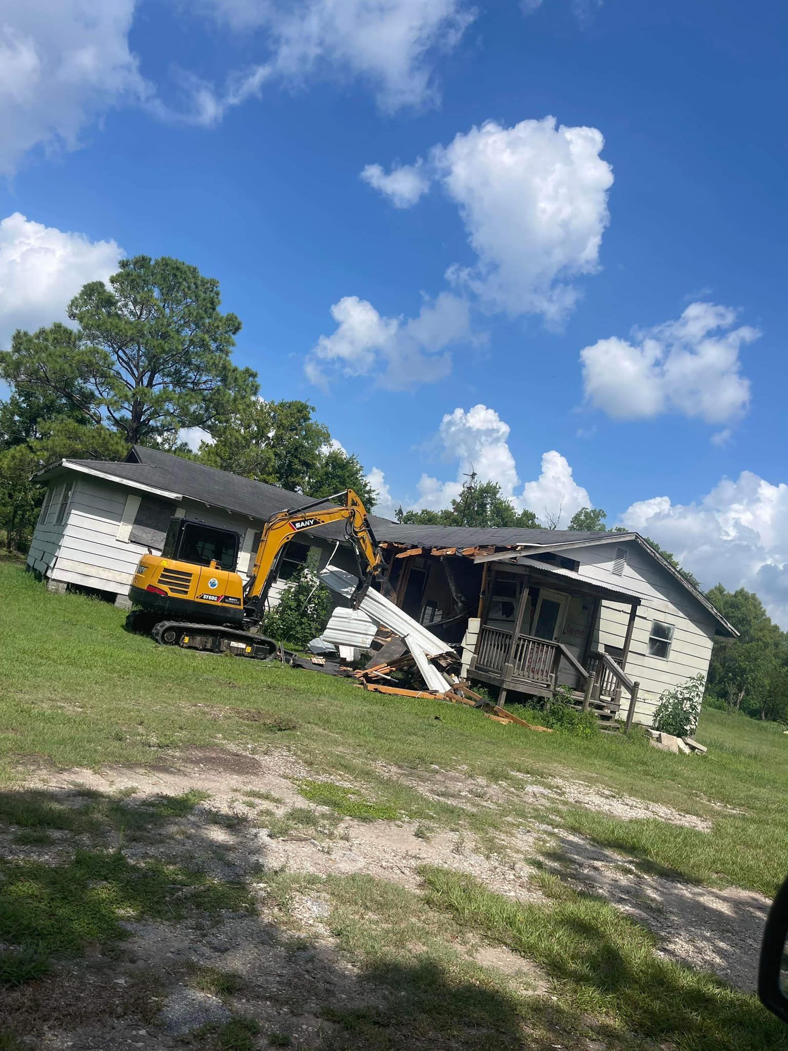 Yellow excavator demolishing a house on a grassy hill under a blue sky with clouds.