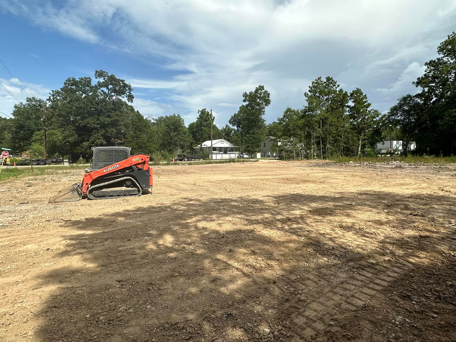 Orange skid steer on a cleared, gravel lot under a cloudy sky. Trees and a house in the background.