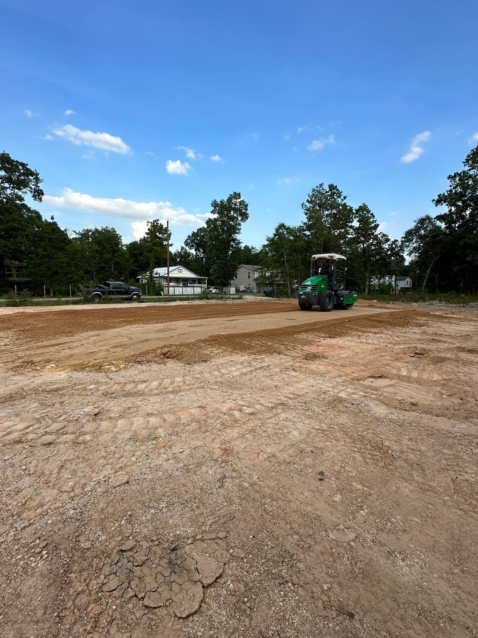 Green construction roller compacts soil on a sunny day with trees and a house in the background.