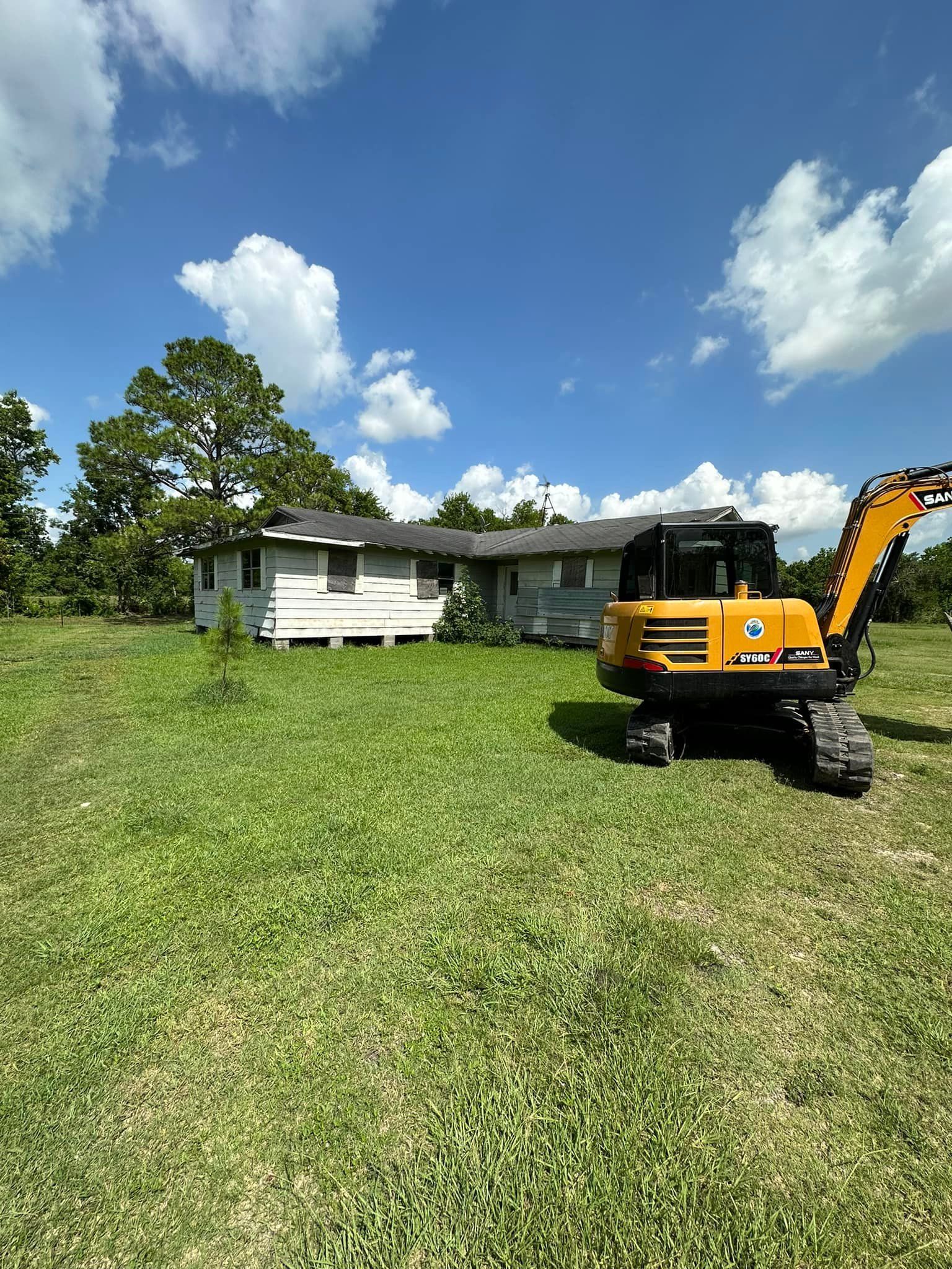 A small, dilapidated house sits in a grassy field, with an orange excavator beside it under a blue sky.
