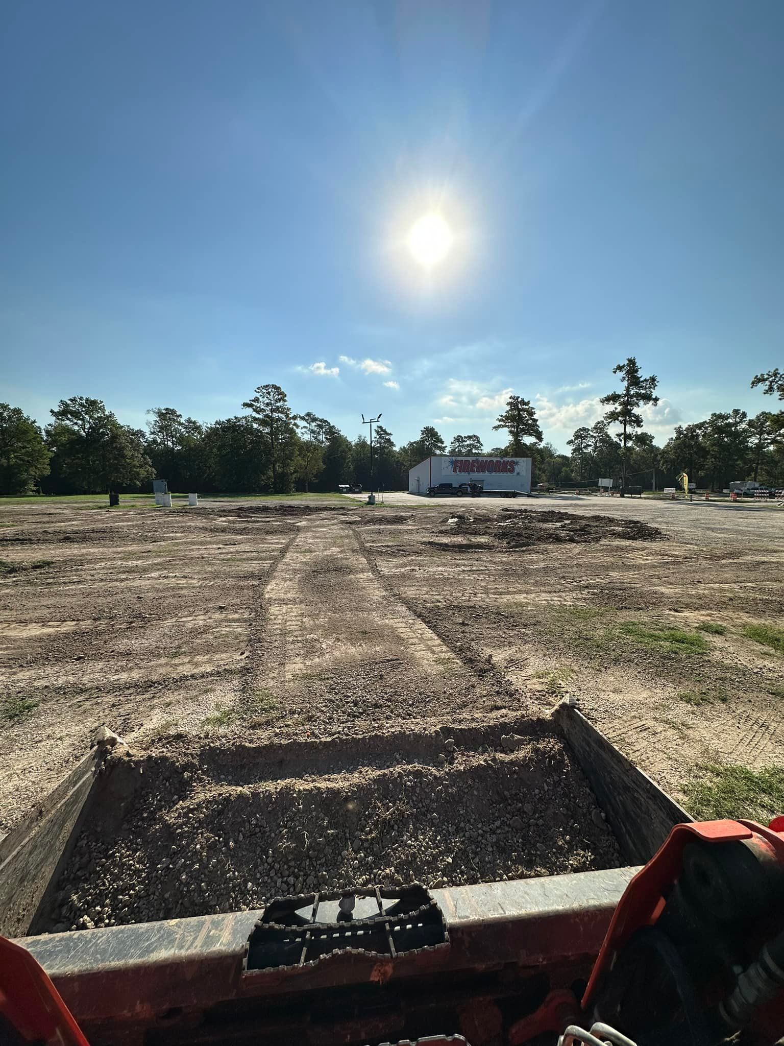 A sunny day over a construction site. A front-end loader bucket is in the foreground, and a building is in the distance.