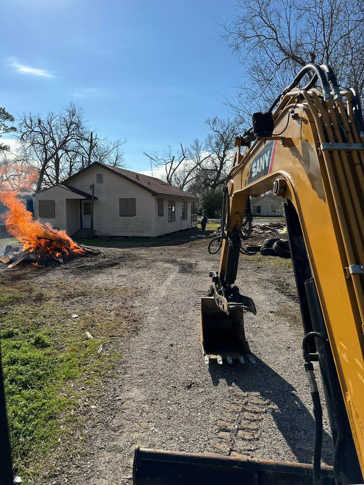 A small excavator sits in front of a house. Fire burns on the left. Bright blue sky.