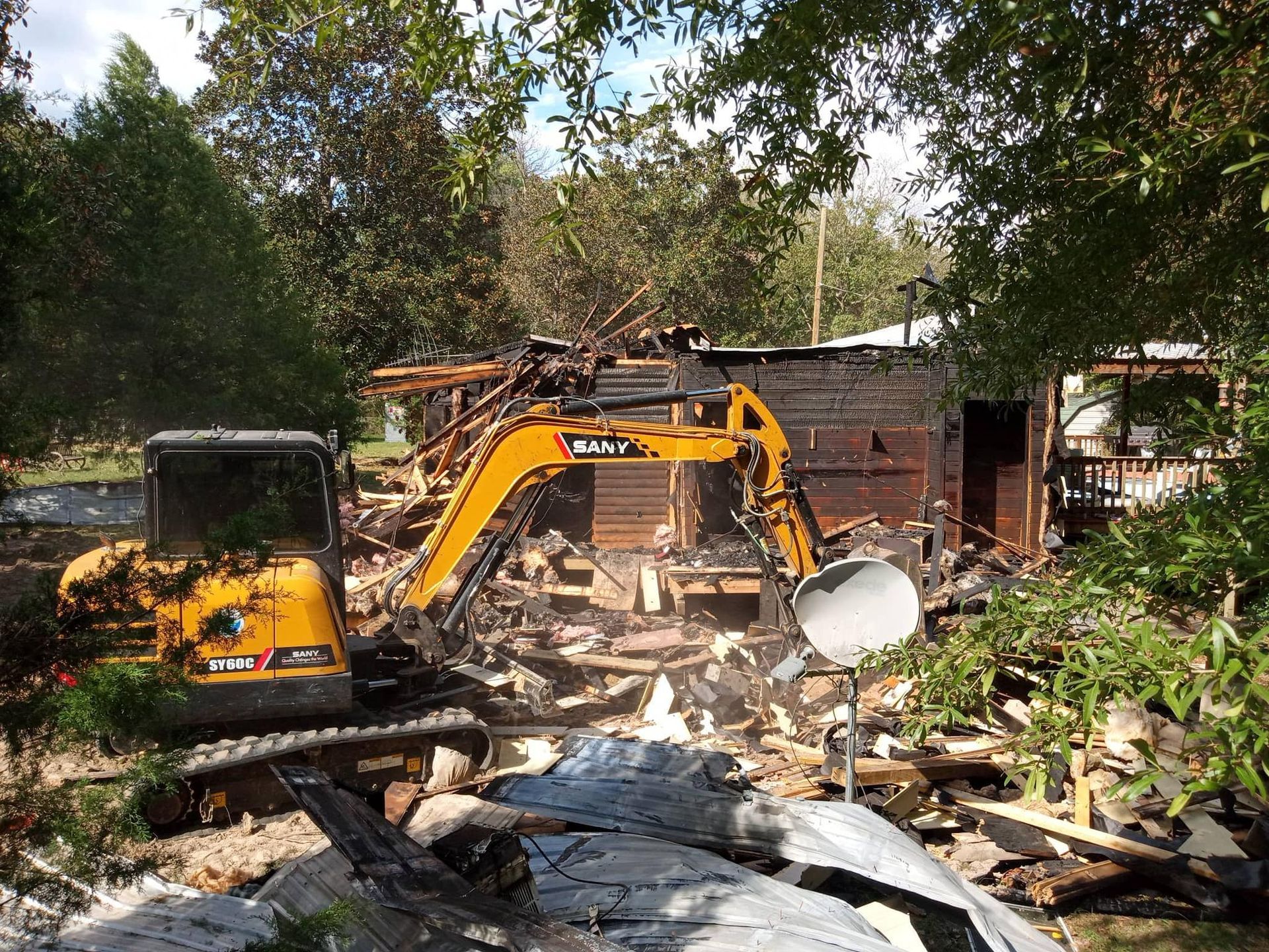 Yellow excavator demolishing a wooden structure amidst debris and trees.