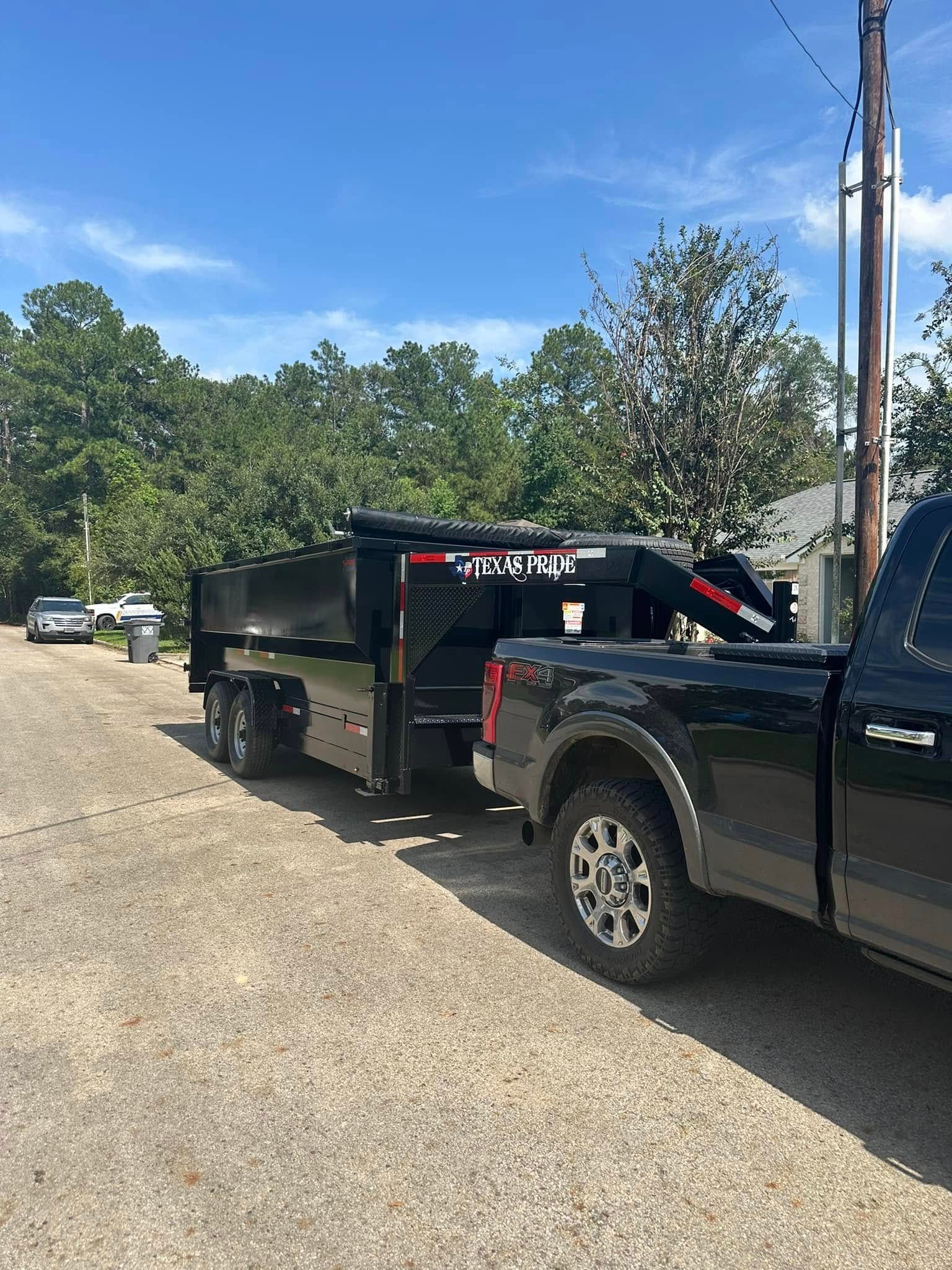 Black pickup truck towing a black dump trailer on a gravel road, trees and blue sky background.