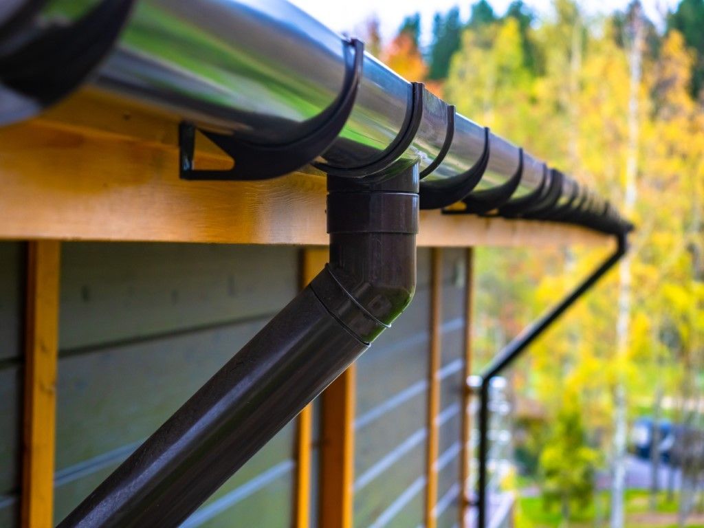 Black rain gutter and downspout on a wooden roof edge with autumn trees in the background