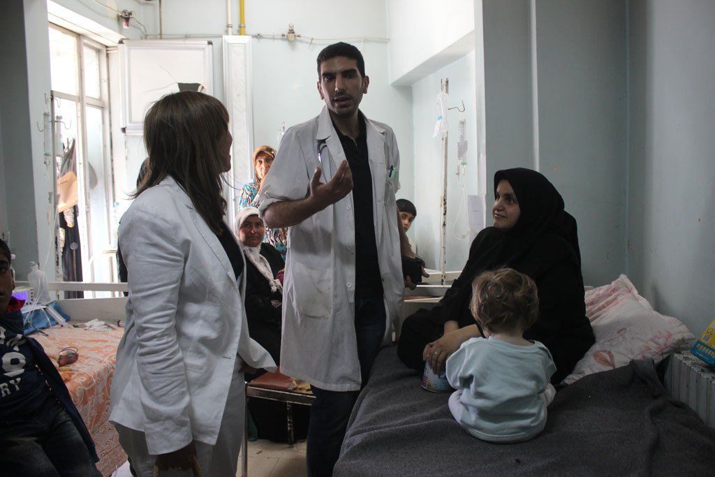 Two Doctors talking with a patient as her child plays and others look on.
