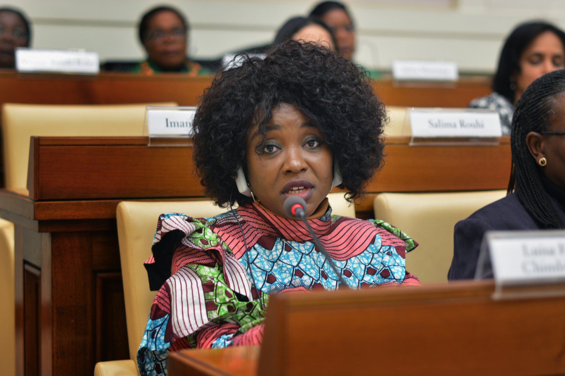 A woman in a colorful dress is sitting at a podium speaking into a microphone.