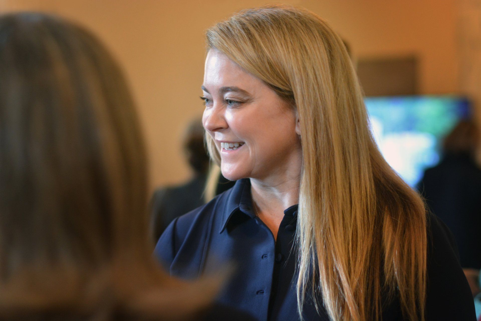A woman with long blonde hair is smiling while talking to another woman.