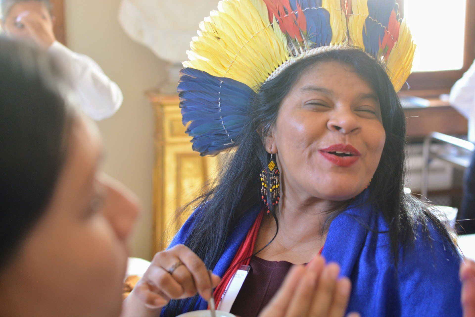 An Indigenous lady wearing a yellow blue and red head dress meeting and talking with others