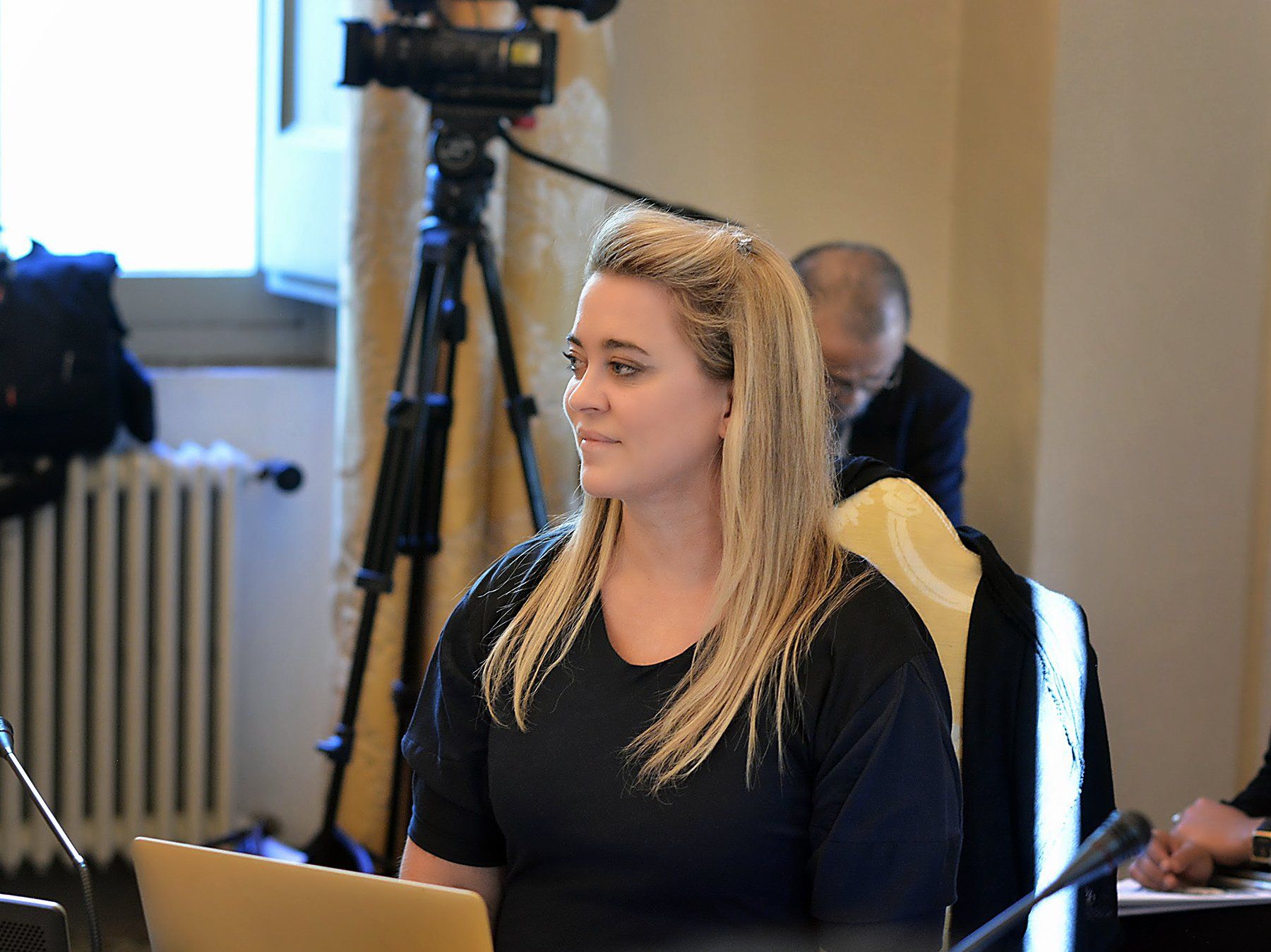 A woman is sitting at a table with a laptop in front of a camera.