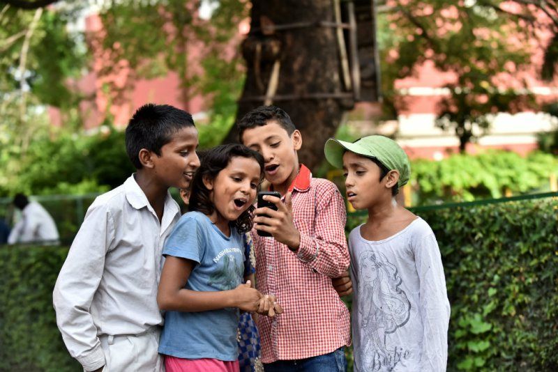 A group of children looking at a cell phone and reacting to what is on it. 