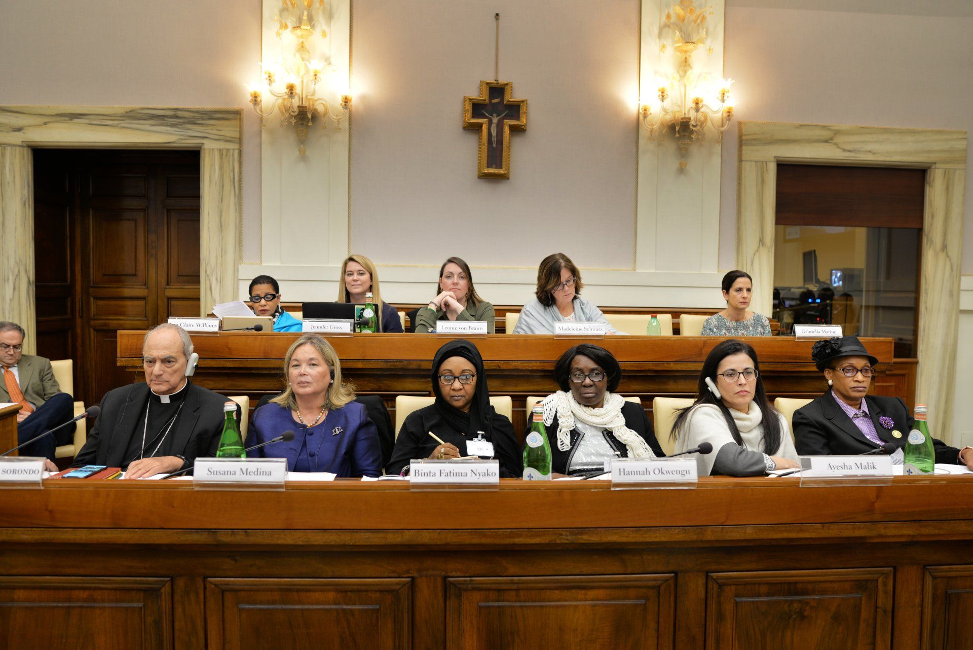 A group of people are sitting at a table in a room with a cross on the wall.