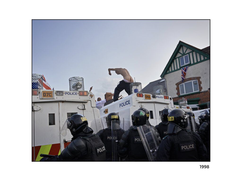 The Peace Project: A man with no shirt , or pants standing on a ambulance surrounded with police in riot gear on the ground