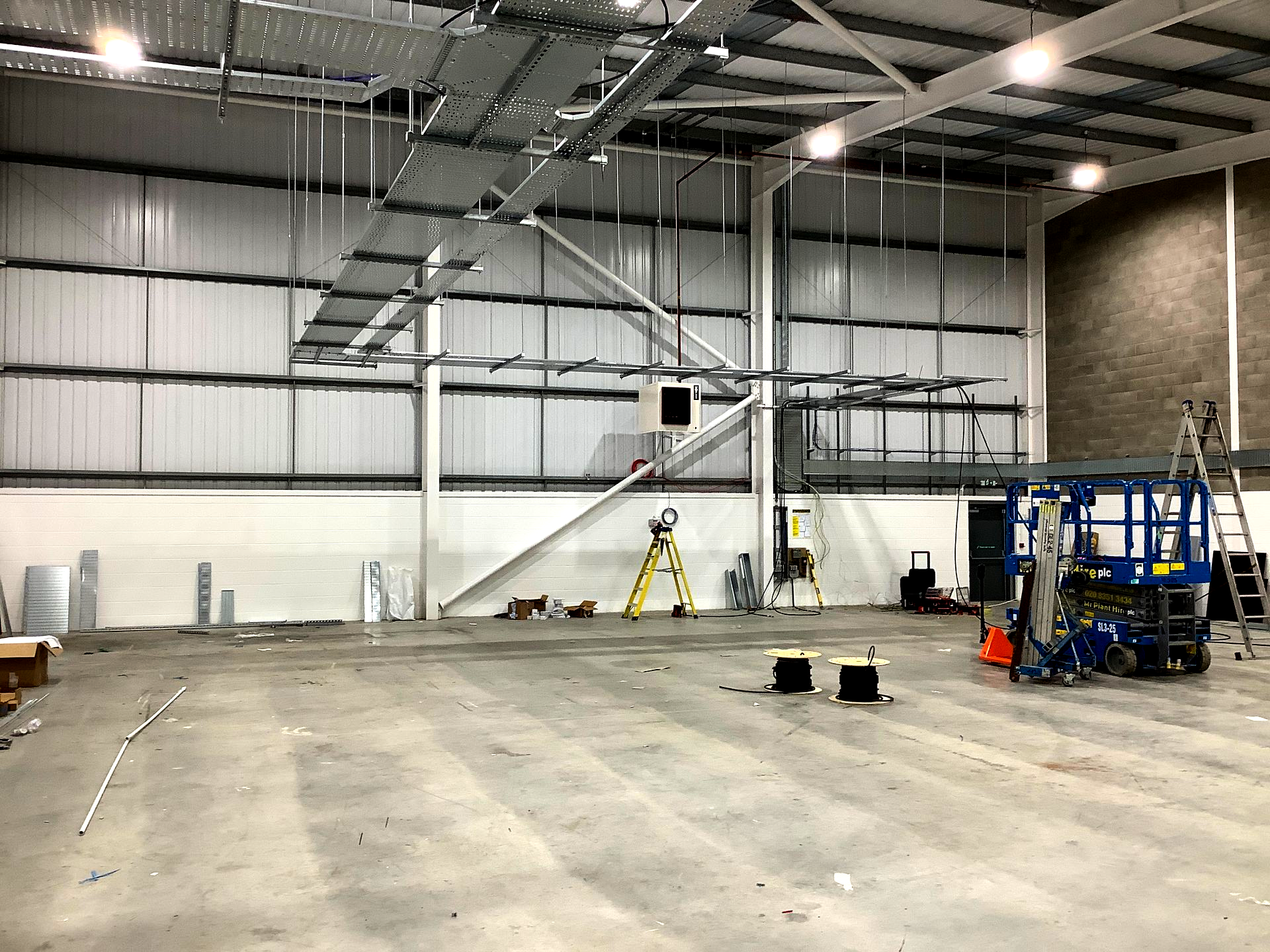 A large, high-ceilinged warehouse interior with overhead industrial cable trays, construction equipment, and tools.