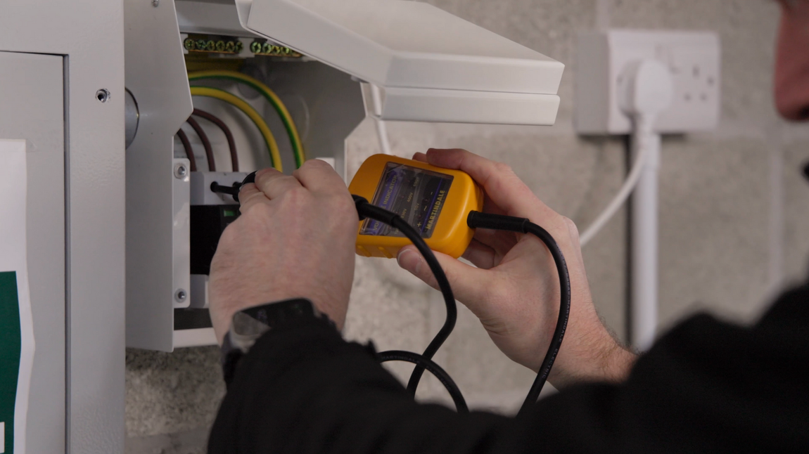 A person uses a yellow multimeter to test electrical wiring inside a wall-mounted panel.