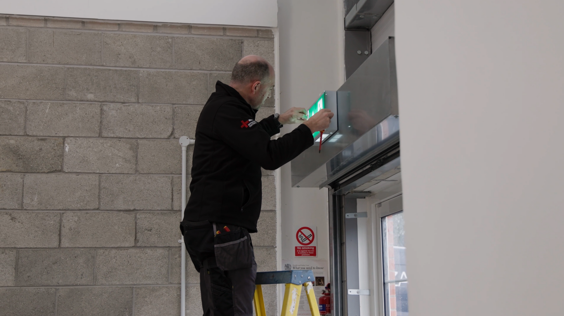 A person on a yellow ladder installs an illuminated green exit sign above a doorway in an industrial building.