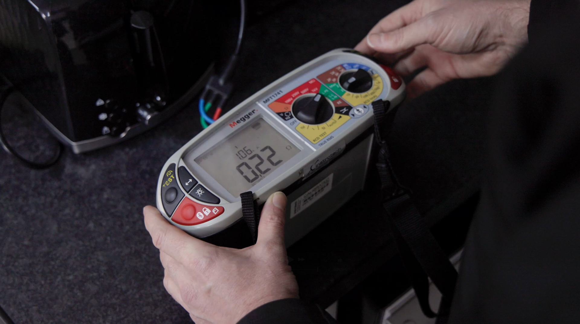 A person's hands holding a Megger electrical test meter displaying a reading of 0.22 near a toaster.
