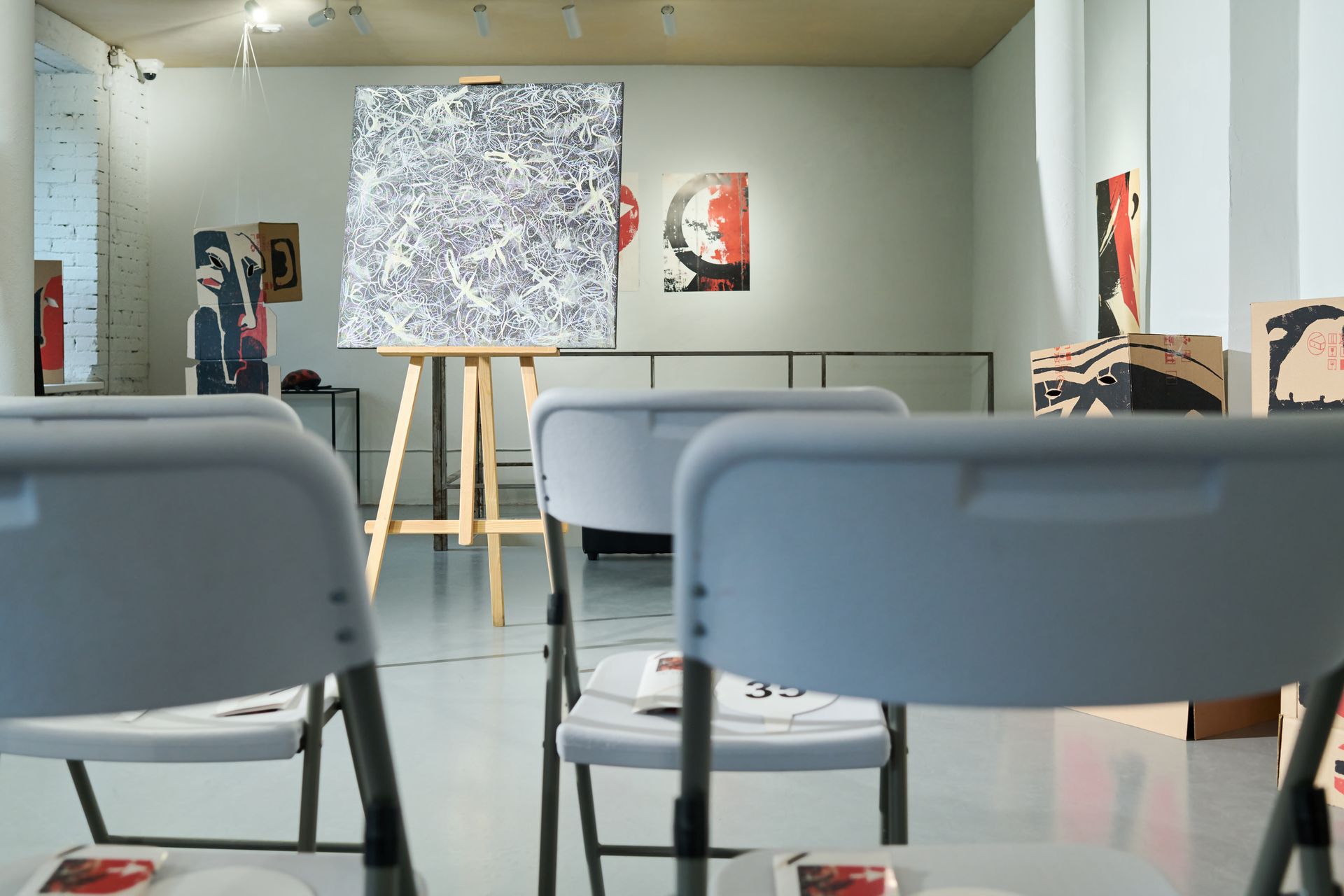 Easel with artwork standing in the center of a fine art gallery in front of white plastic chairs. Easel with artwork standing in the center of a fine art gallery in front of white plastic chairs.