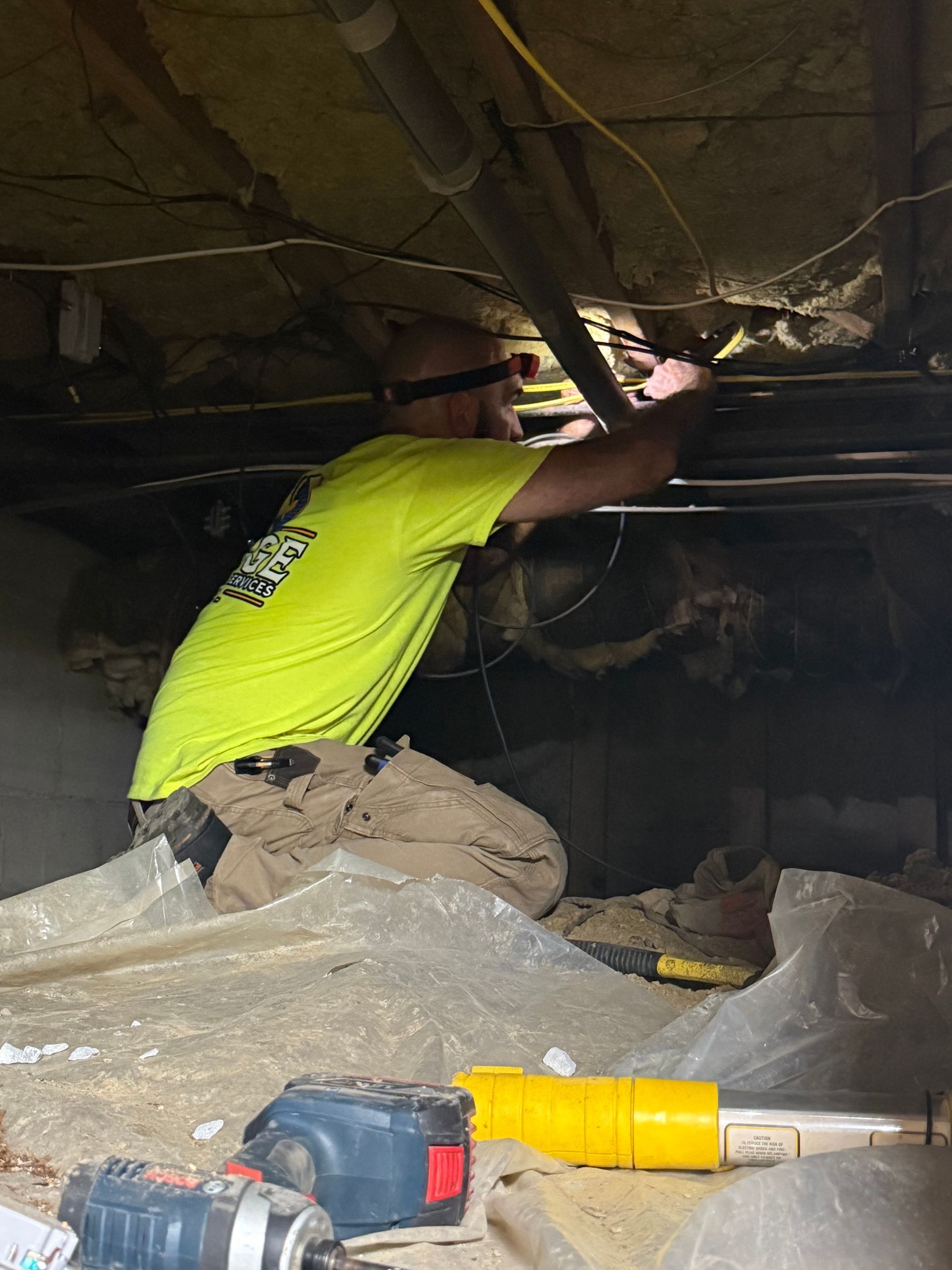 A person in a neon yellow shirt works beneath pipes in a crawl space. They're using a light, tools are in the foreground.