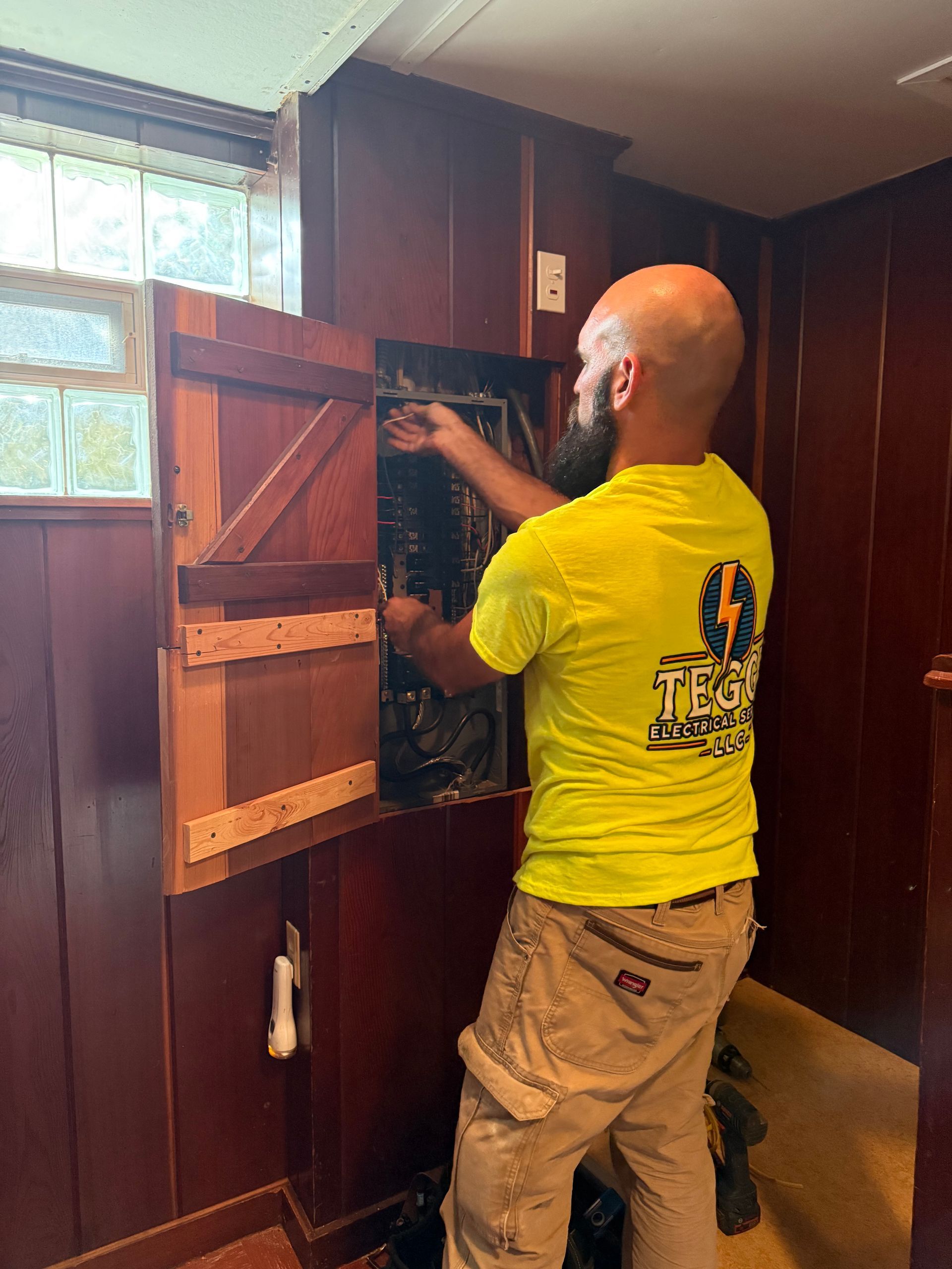 An electrician in a yellow shirt works inside an electrical panel. He stands in a room with wood-paneled walls.