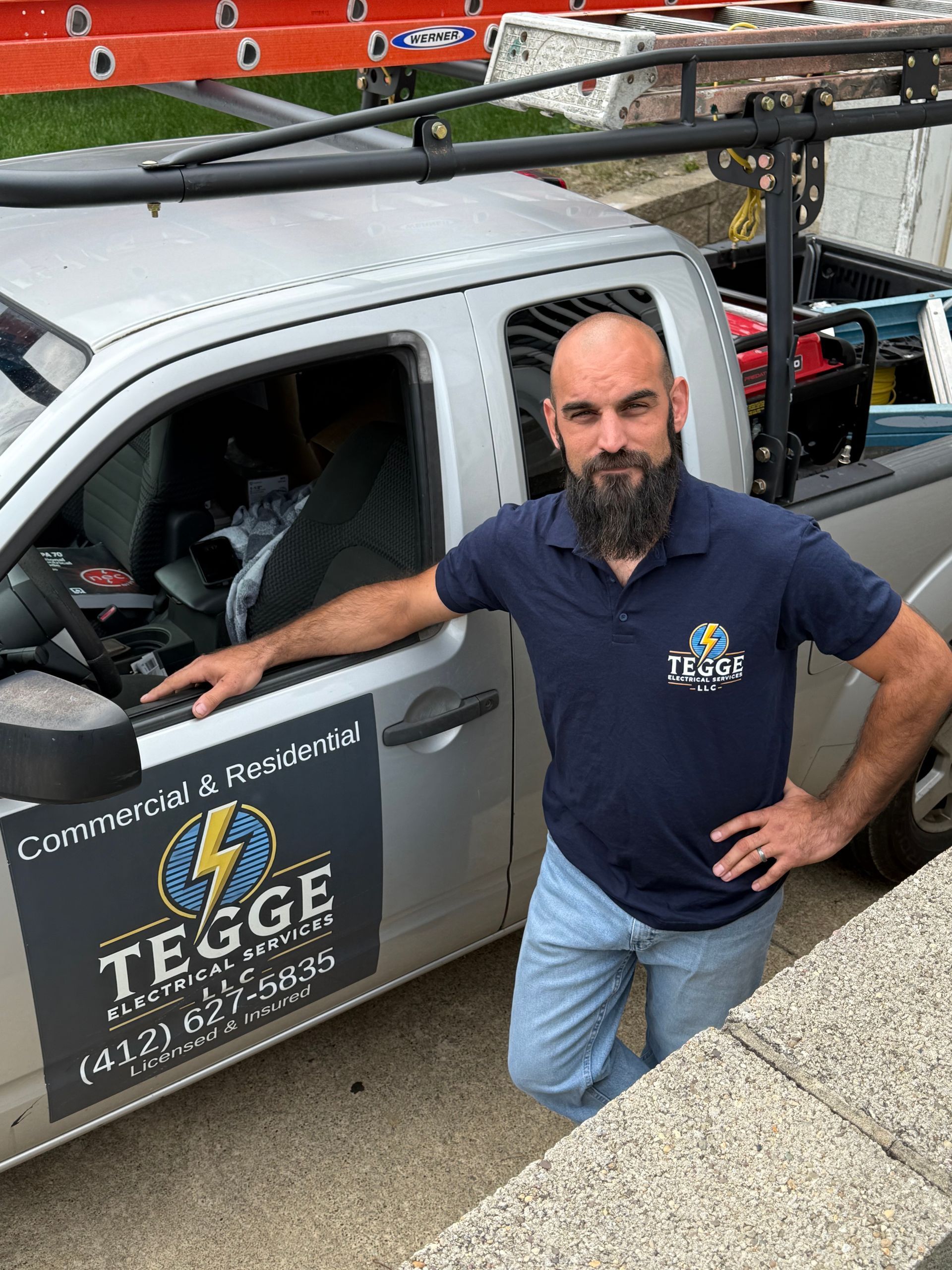 A man with a beard stands next to his work truck with the logo