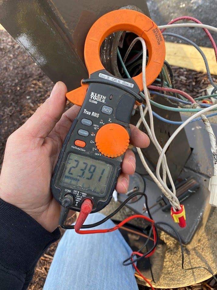 A hand holding a multimeter measuring amperage on an outdoor HVAC unit; reads 23.91.