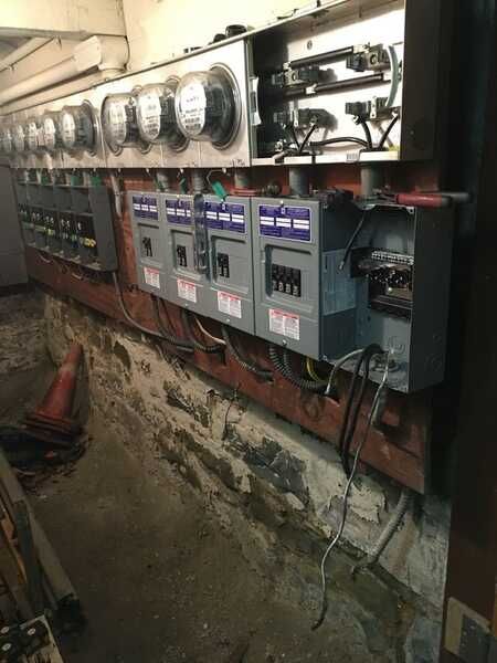 An electrician in a yellow shirt works inside an electrical panel. He stands in a room with wood-paneled walls.