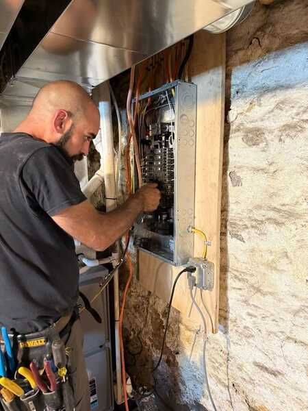 A man with a beard in a yellow shirt crouches in a basement, looking into a tool bag. He is working on electrical wiring.