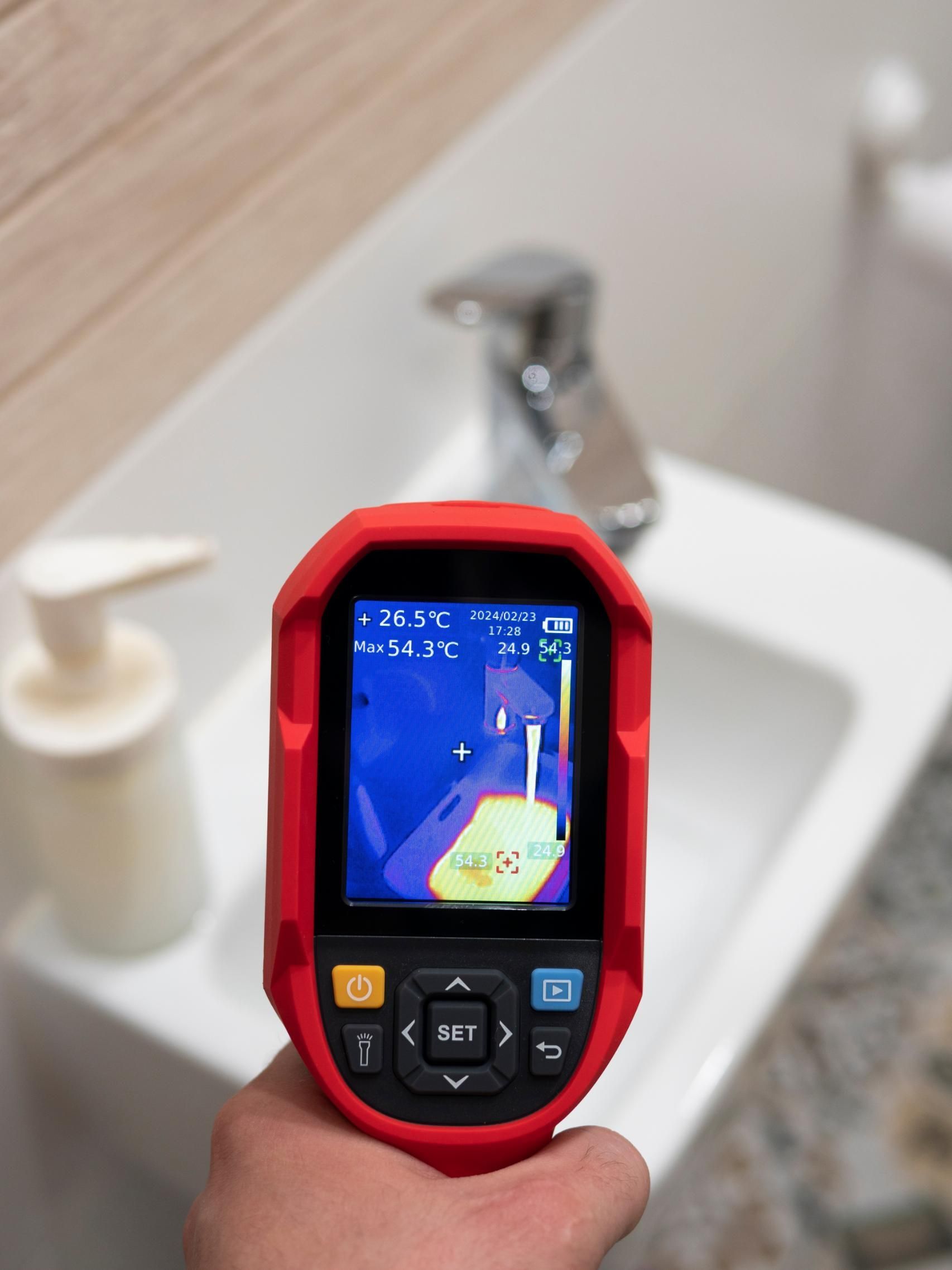 Hand holding a red thermal camera pointed at a bathroom sink, displaying temperature readings.