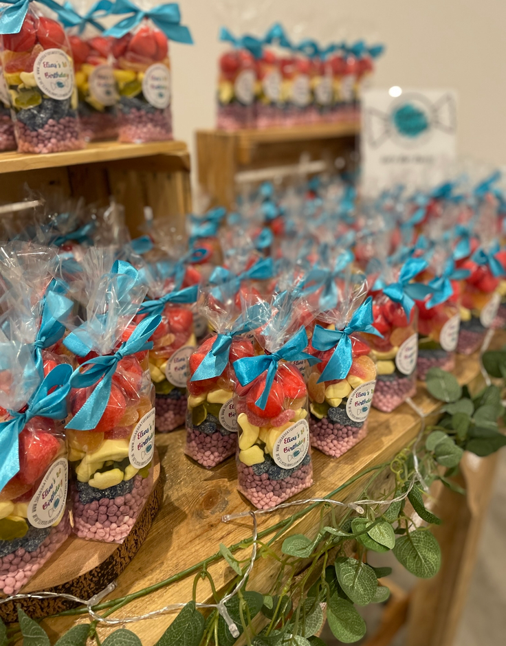 Candy favours in clear bags tied with blue ribbons, arranged on wooden shelves.