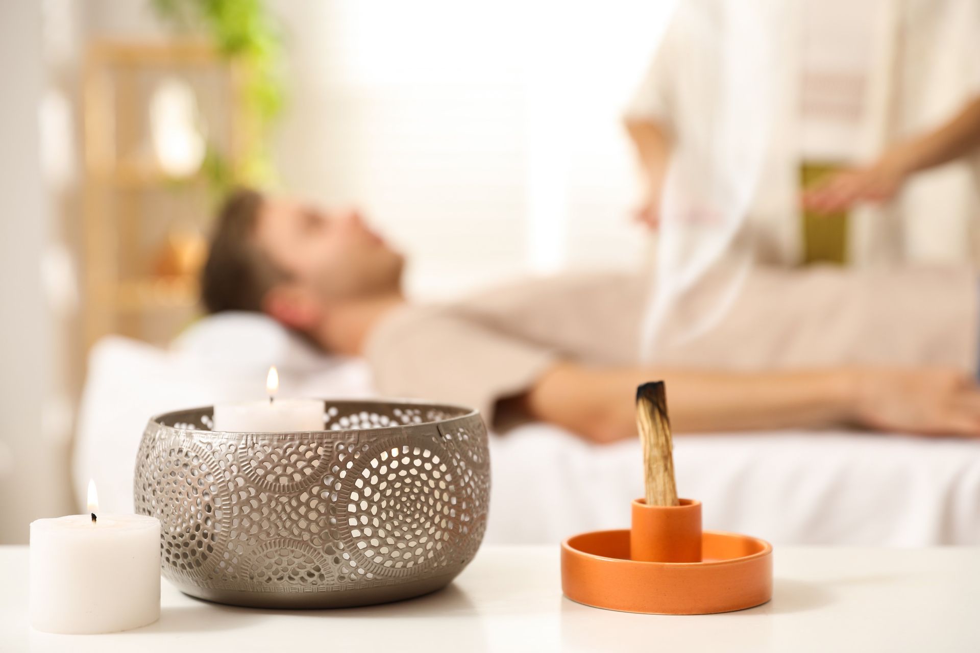 A spa setup with a candle, incense holder, and decorative bowl in front of a blurred reclining person.