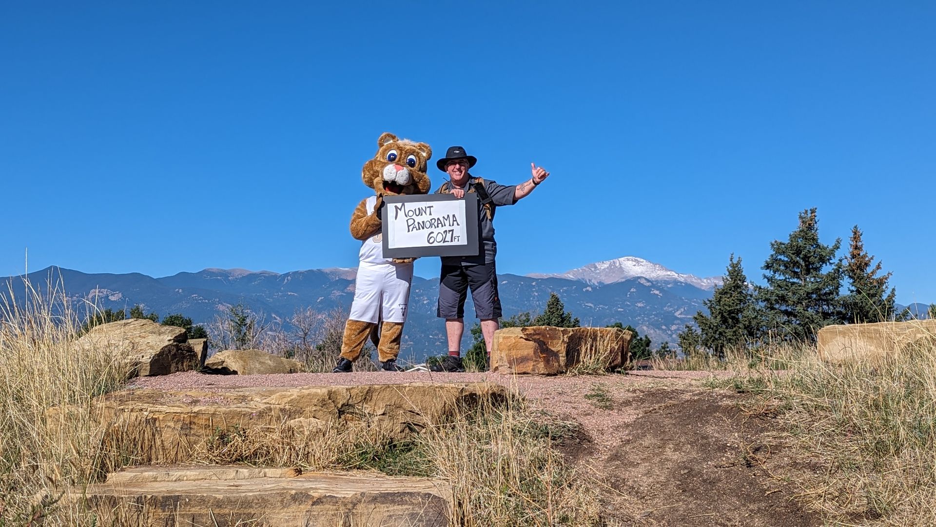 A man and a person in a mascot costume with a sign that says Mount Panorama 6027 feet stand on top of a hill with Pikes Peak in the background