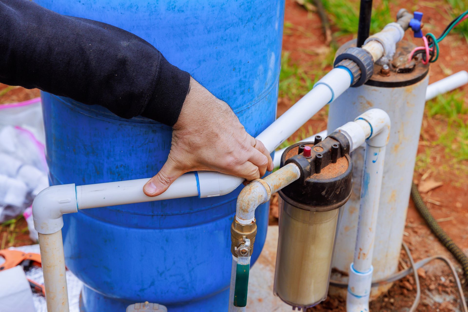 A person's hand stabilizes a white PVC pipe connected to a blue water tank and a filtration system outdoors.
