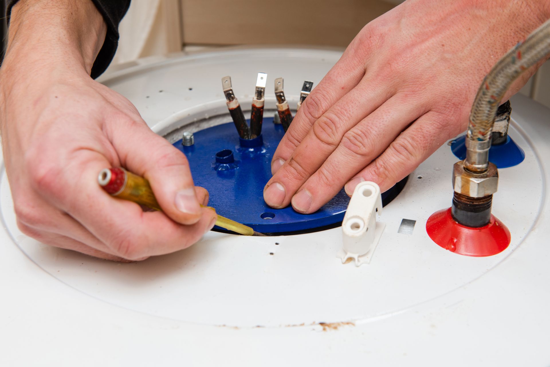 Hands working on a water heater, using a tool to tighten something near electrical terminals.