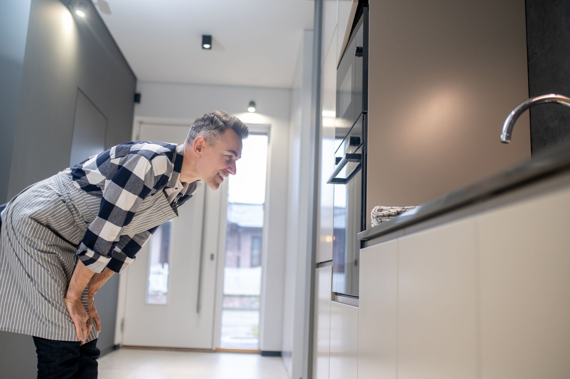 Man in apron looking into oven in kitchen.