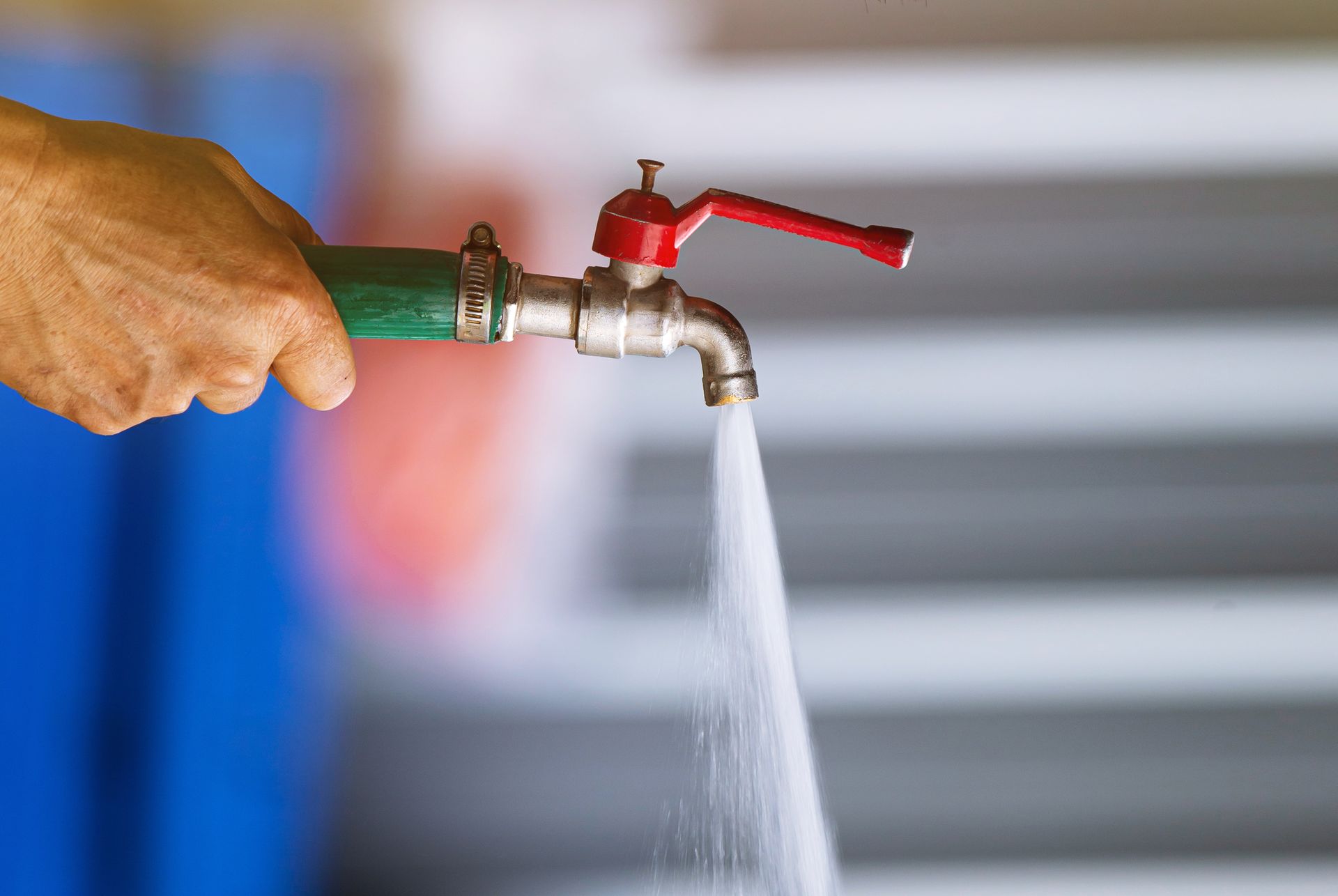 Hand holding a water faucet with water flowing out. The faucet is silver and red; the hose is green.