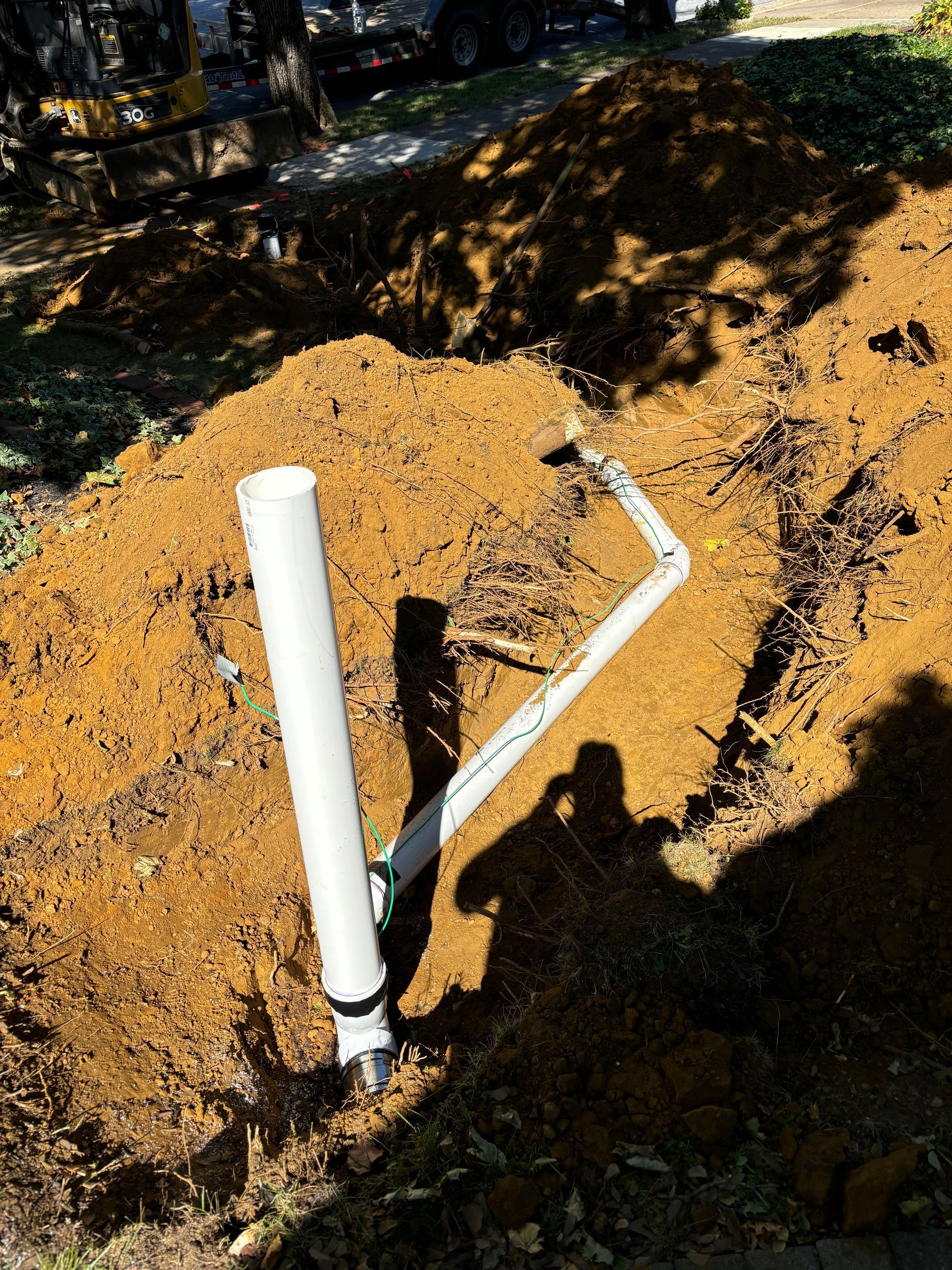 White PVC pipe and angled metal pipe exposed in a dirt hole. Sunlit outdoor setting.