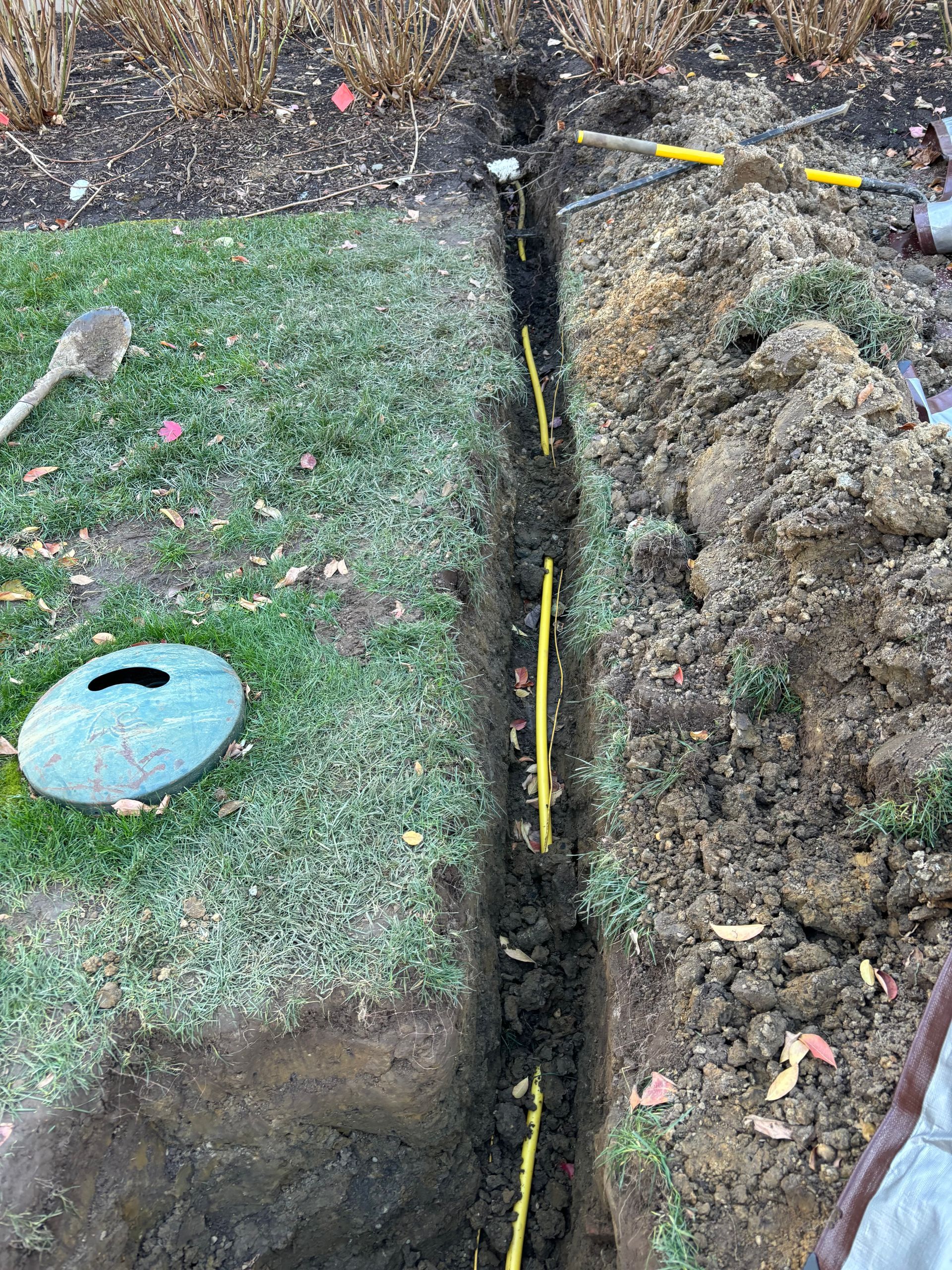Trench with yellow pipes in grass and dirt. Tools and a green lid are nearby.