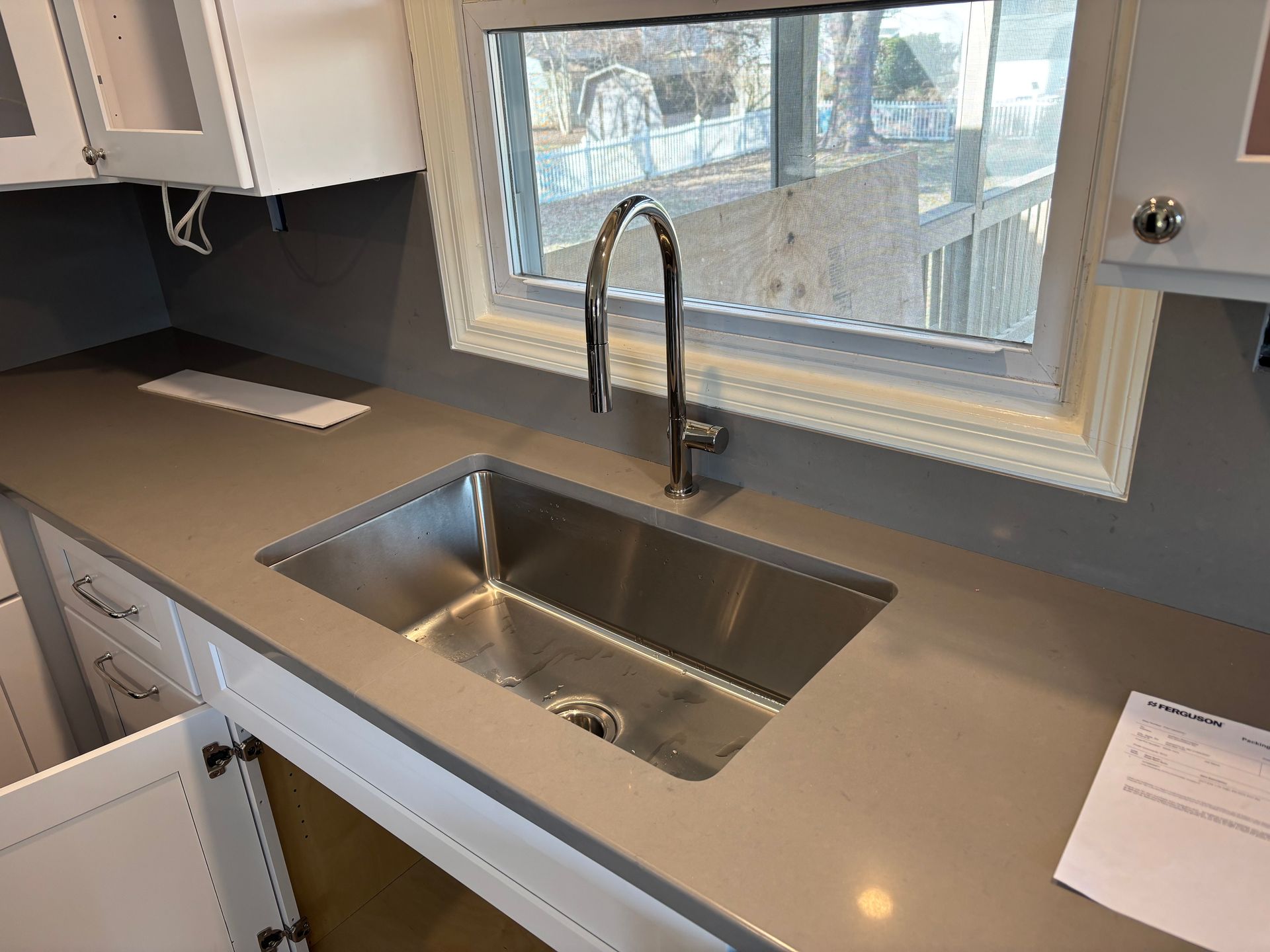 Kitchen sink in a gray countertop, with a window behind it. White cabinets are visible.