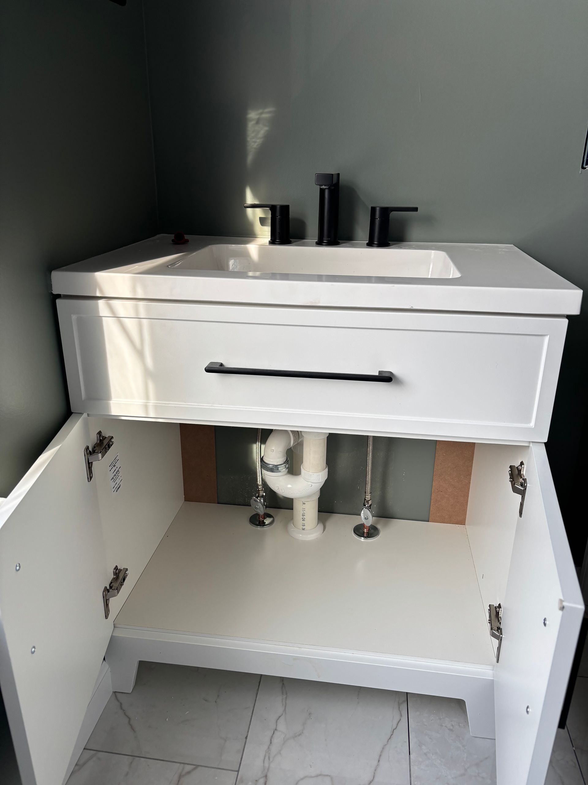 White bathroom vanity with open doors, black fixtures, and plumbing visible.